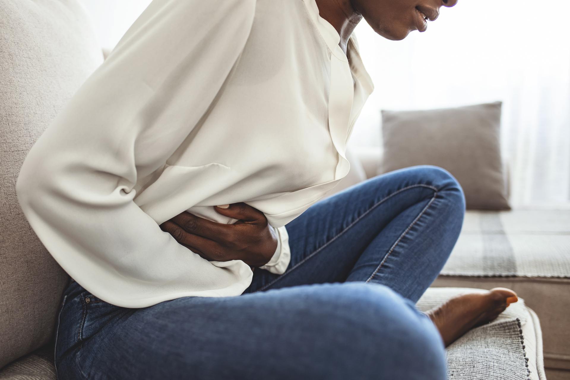 A woman holding her stomach in pain wearing an off-white blouse and blue jeans sitting on the couch.