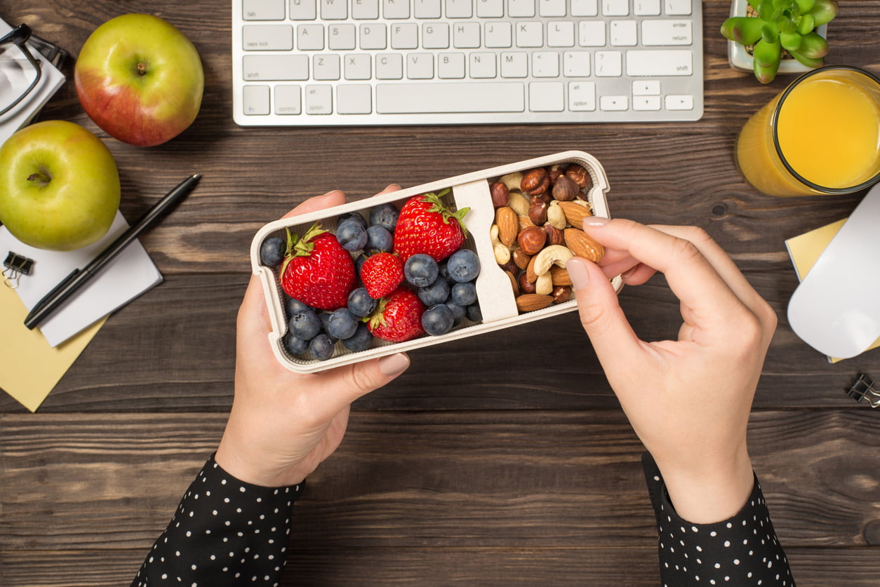 A woman’s hands holding a lunchbox with nuts, berries, and an apple on an isolated dark wooden table