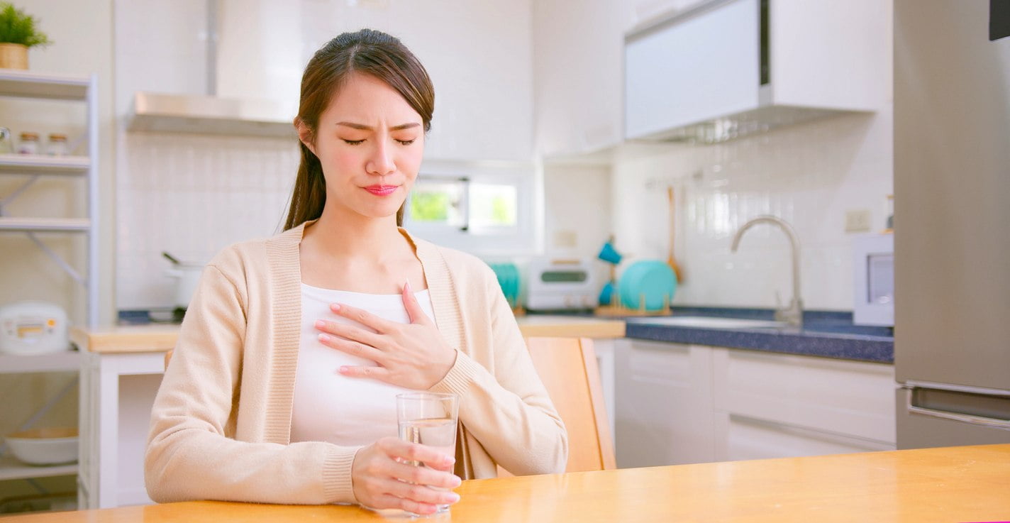 A brunette woman in cream colored shirt sitting at a table with her hand over her chest expressing discomfort.