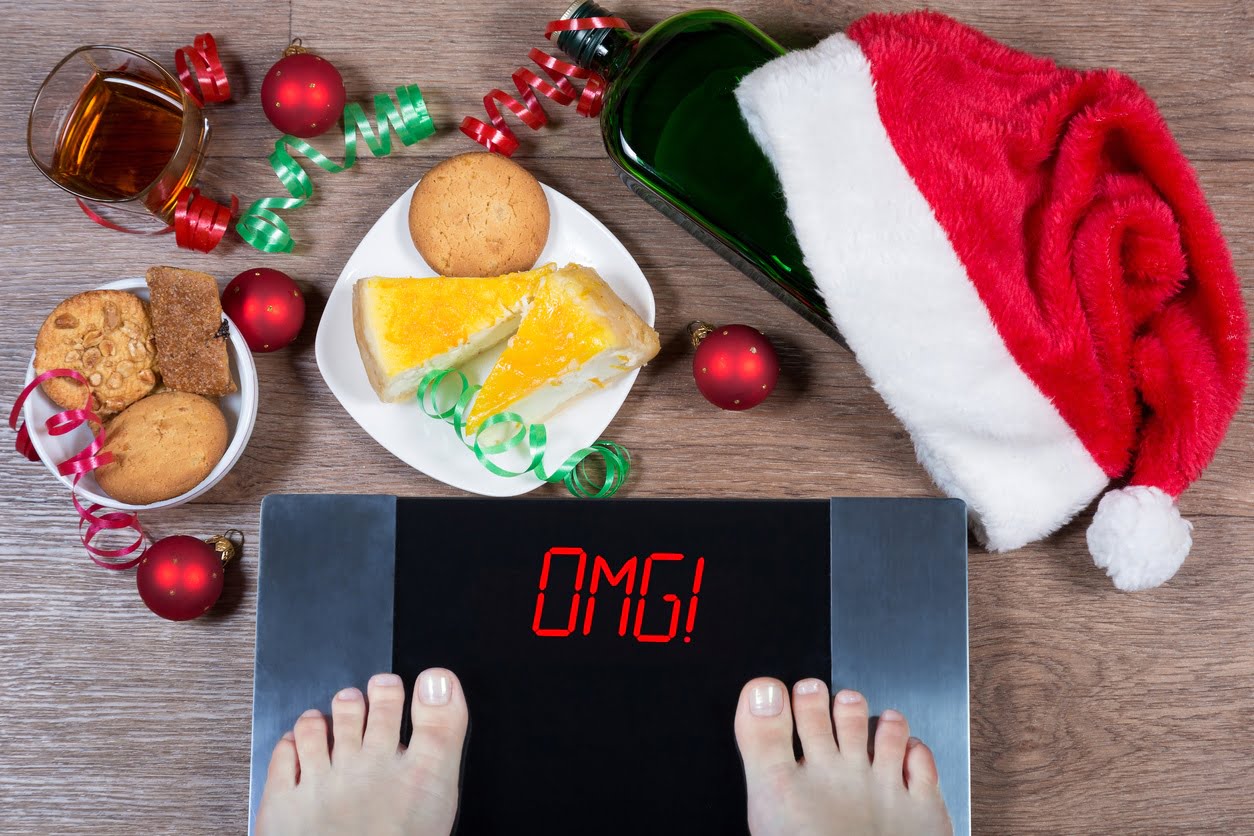 Female feet on digital scales with a sign "omg!" surrounded by Christmas decorations, bottles, a glass of alcohol, and sweets.