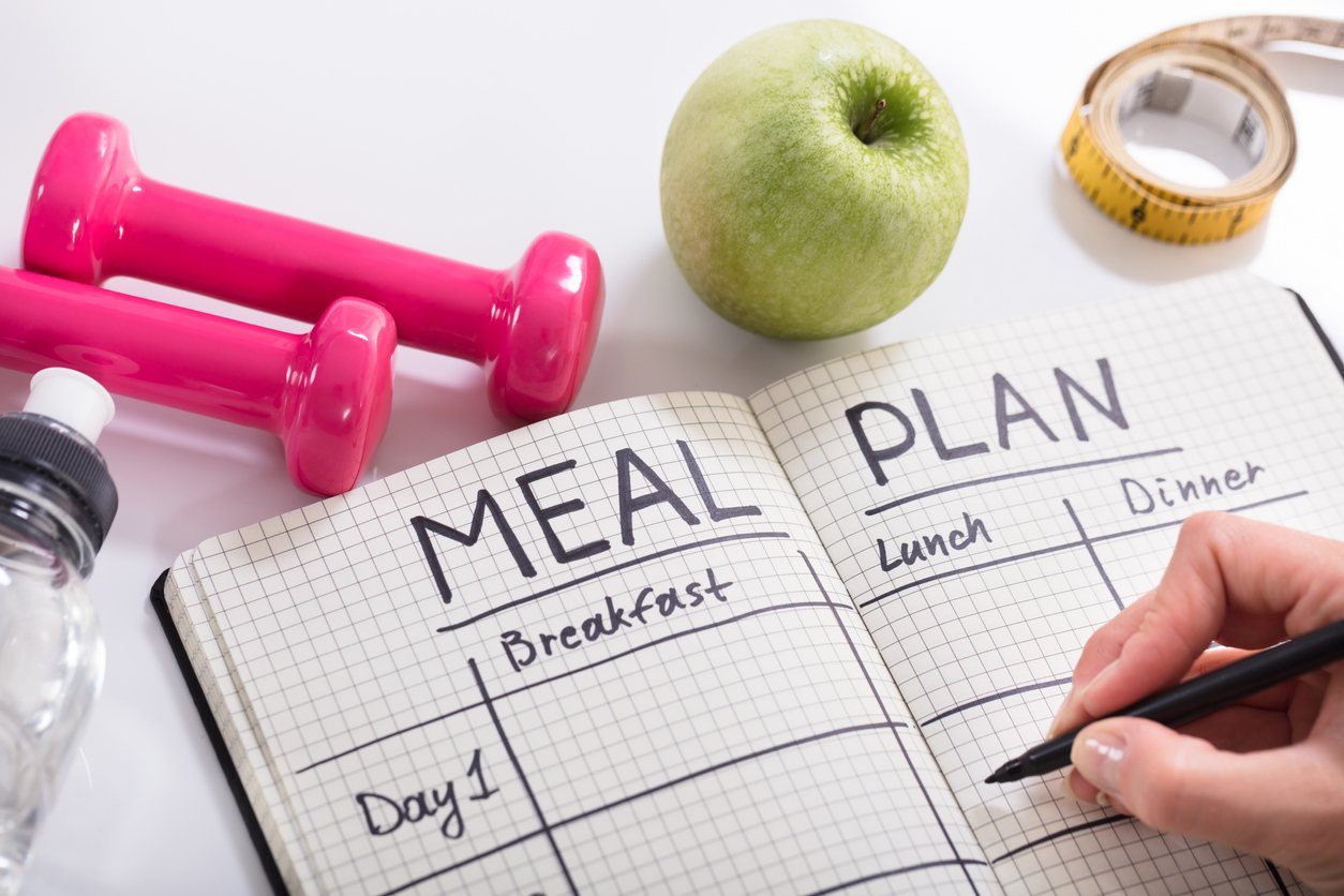 A woman filling out a meal plan on a wooden desk near a green apple and pink hand weights.