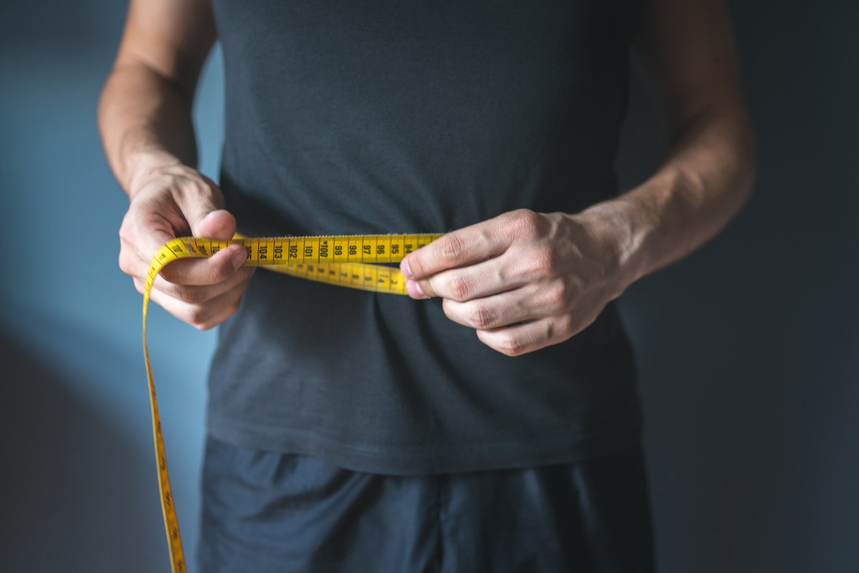 A man in a grey shirt measuring his waist line after weight loss.