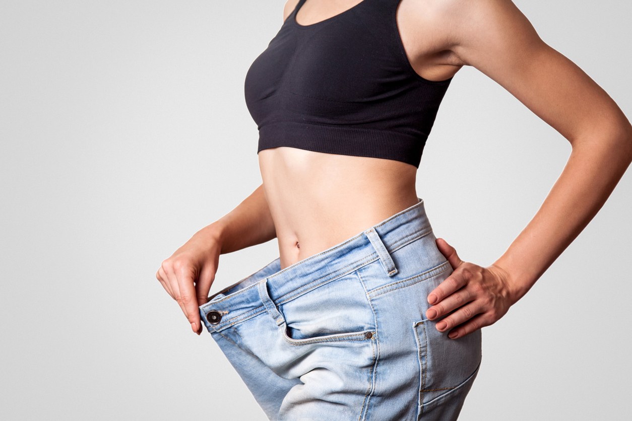 A woman in a black top and blue jeans holding out the jeans' waistband illustrating weight loss.