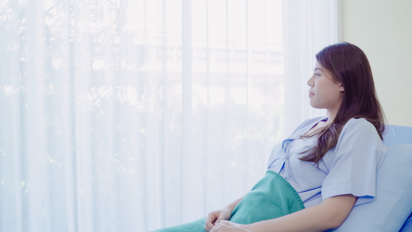 An adult woman in blue scrubs and a green blanket resting in a hospital bed recovering from gastric sleeve surgery