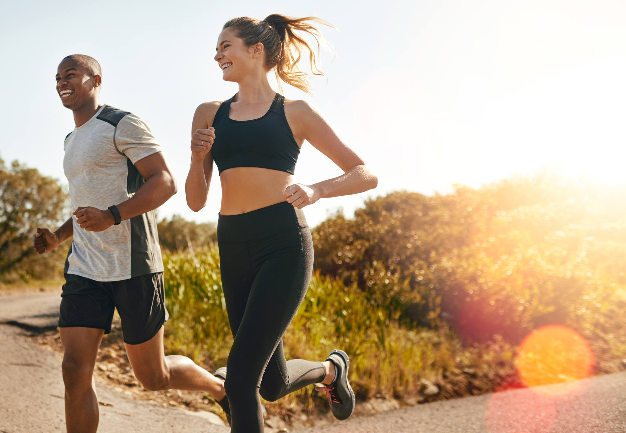 A man and woman in sports clothes are smiling as they run down a path with the sun behind them.