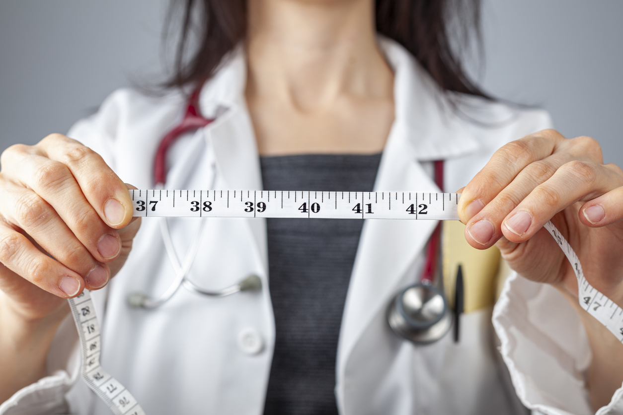 A doctor wears a white coat and a stethoscope while holding up a tape measure to represent weight loss from bariatric surgery.