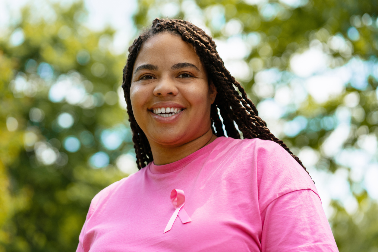 A woman with a bright pink shirt and breast cancer awareness ribbon smiling brightly. 